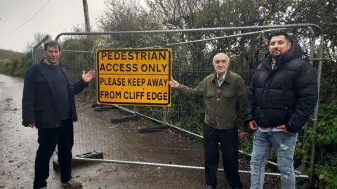 Somerset Council Three men stood on a cliff-top road. It has a big sign saying "Pedestrian Access Only - Please Keep Away from Cliff Edge"