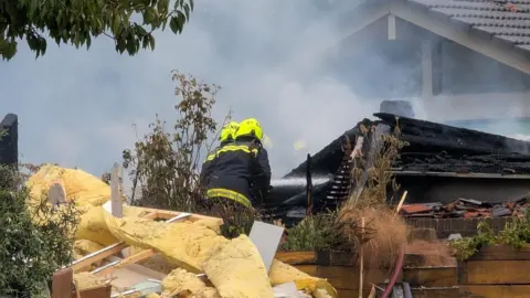 Two firefighters with neon yellow hard hats on and black and yellow outfits. They are among a pile of rubble, surrounded by smoke which is rising. 