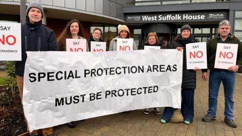 A group of seven protesters – men and women – standing outside a council building. They are holding up signs which read "Say NO to Cranswick" and a large banner which reads "Special Protection Areas Must Be Protected".