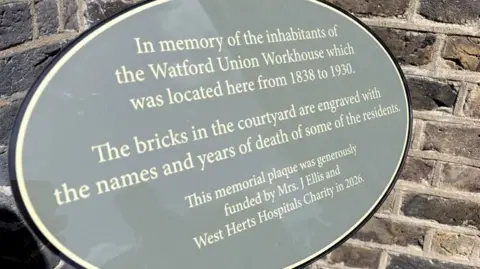 A close up of the grey, circular plaque that remembers the people that were housed at the workhouse. It says it's funded by a former patient of Watford General, Mrs J Ellis. 