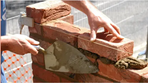 Close up of hands laying bricks with a trowel. There is a building site in the background.
