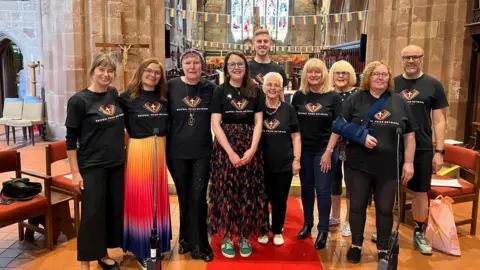 Ian Handley A group of people are inside a church. They are dressed in black T-shirts with a rainbow and a cross on them. Behind them, rainbow flags are strung up on banners.