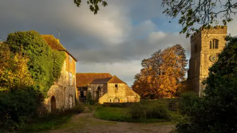 A medieval palace. The palace has stone walls and a tiled roof. The building is surrounded by trees and other buildings. The scene is at sunset.