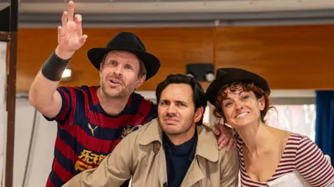 Michael Berkeley A close-up of three actors, one female, in rehearsal for The 39 Steps, two are wearing black trilbys and t-shirts, the other one is in a beige trench coat. 