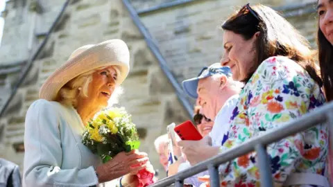 Reuters Queen Camilla is seen on the left of the image, facing towards the right. She is wearing a bright blue suit and a beige hat. She is holding a small bunch of yellow flowers. Camilla is speaking to someone in the crowd.