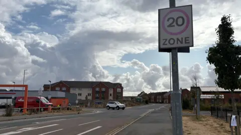 A white car turning out of a junction of a new build estate, with a close up of a 20mph speed sign next to it. 