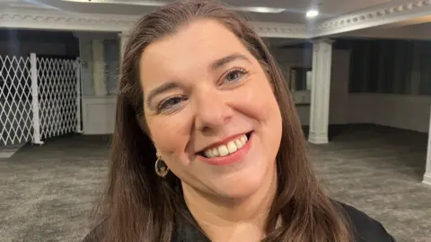 Elaine smiling at the camera. She has long brown hair and is wearing a black top and gold hoops.