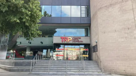 The exterior of the Theatre Royal Plymouth. There are steps leading up to the buildings which has glass doors which are reflecting the building opposite. There is a tree to the side.