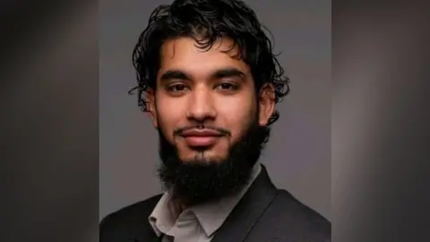 Bedfordshire Police Adam Khan: a man who is sitting looking at the camera and smiling. He has black hair and black facial hair. He is wearing a grey suit jacket and light grey shirt.
