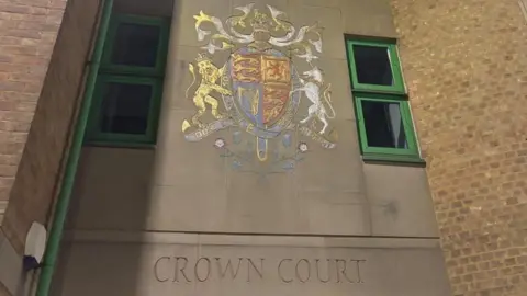 Brian Farmer/BBC Luton Crown Court: The words "CROWN COURT" written on a grey wall below a multi-coloured crest. Two window with green frames and brown-brick walls are also visible at the sides of the crest.