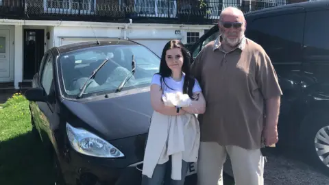 A teenage girl with dark hair standing next to an older man with a white beard and sunglasses on. They are standing in front of a black car which is parked in front of a house.