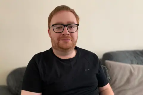 A man with ginger hair and a beard in a black t shirt stares into the camera