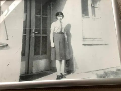 Handout A black and white image of Sandy Cawley. She is wearing a beret, tie, skirt and white shirt. She is standing in front of a doorway. 