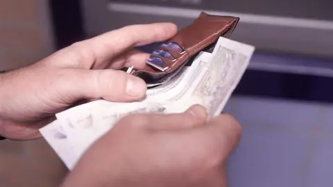 A man slides banknotes into a brown leather wallet.