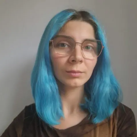 Vlada Bondar A young woman with bright blue shoulder-length hair and large square glasses looks into the camera. She wears a long-sleeved brown t-shirt.