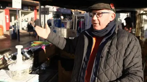 A man in a quilted black jacket, flat cap, black snood and orange jumper. He is standing at a stall which is selling lighting and other miscellaneous items.