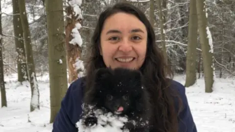 family handout Maria holds a little black fluffy dog and smiles at the camera. They are standing in woods that are covered in snow, and the dog has snow on its tummy. Maria wears a blue fleece and has very long black wavy hair.