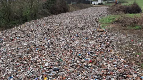 A field that is completely covered in rubble and mixed waste, including broken bricks, stones and bits of plastic. Bushes and fields line the sides.