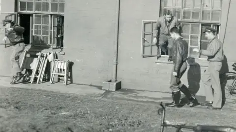 IWM Duxford A black and white photo of four US airmen at a building with two large windows at RAF Duxford during World War Two. One airman has just jumped out of the window on the left. On the right, an airman is half in and half out of a window, ready to leap. The two other airman are walking on the pavement ahead of him. 