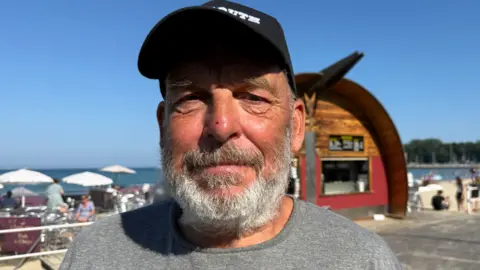 A man wearing a cap and a grey t-shirt looking into the camera on a sunny day next to the beach