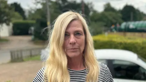 Elliot Deady/BBC Jackie Bendon standing outside her home with a sewage tanker in the background