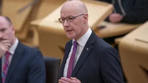 PA Media First Minister John Swinney is mid speech in the Holyrood Chamber. He is bald with glasses and is wearing a navy suit which has a saltire badge on it. He also has on a white shirt and purple tie.