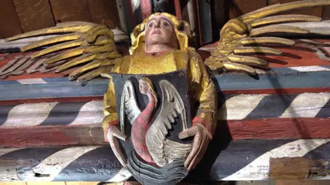 A carved and painted wooden angel, wings outstretched and painted gold. It holds a shield with a woman stylised to resemble a bird. It is attached to a well-decorated beam with red and barber pole style stripes.