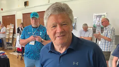 Warren Muggleton/BBC Steve Perryman smiles at the camera as if about to speak. He wears a navy blue jersey shirt with an open collar and one button done up He is clean shaven and has grey hair. Behind him a man in a blue baseball cap with a lanyard round his neck also looks at the camera. In the background, men mill about the stalls.