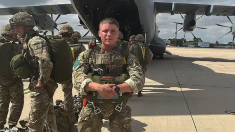 Ashley stands in a wide stance looking at the camera. He is dressed in camouflage gear and has a parachute strapped to his body. Behind him a plane is parked and soldiers are boarding the plane behind him on the runway. 