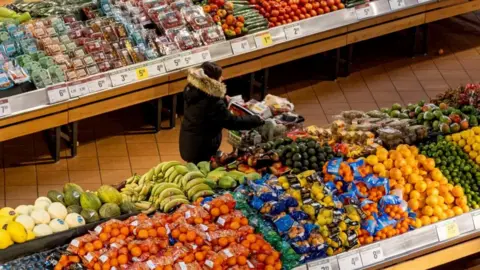 A woman in a dark coat and seen from above pushes a shopping cart through the produce section of a grocery store. On either side of her are piles of brightly coloured produce.