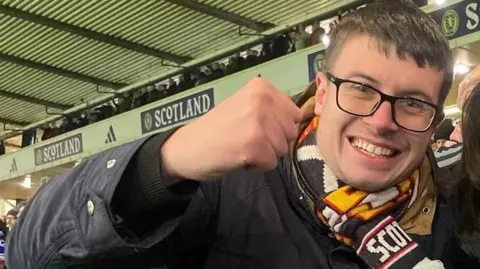 Daris Nesbitt Daris Nesbitt, who is wearing a navy jacket and a Scotland scarf, waves his right fist in the air as he celebrates the national team's 4-2 victory over Denmark at Hampden. He has short dark hair, dark-rimmed glasses and is grinning widely.