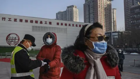 Getty Images A Chinese woman (left) wears a protective mask as she has her temperature checked by an official entering a park on 9 February 2020 in Beijing, China