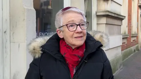 A woman stood in front of an empty shop on a high street. She has grey hair with a purple streak and is wearing a black coat with a fur hood and a red scarf. 