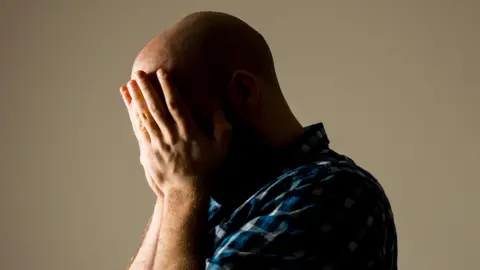 A man wearing a blue checkered shirt and holding his head in his hands.