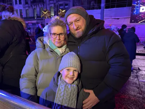 Two parents and a young boy, wearing hats, scarves and jackets, smile at the camera