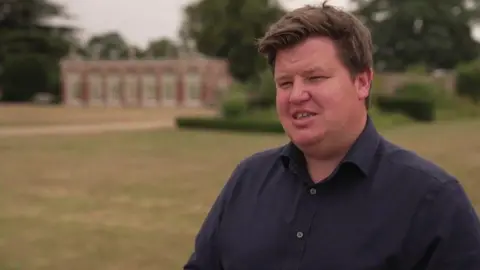 Alasdair Hutson standing outside Burton Constable Hall. He is looking to the left of the camera. He has short, brown hair and is wearing a navy shirt, with the top button undone. 