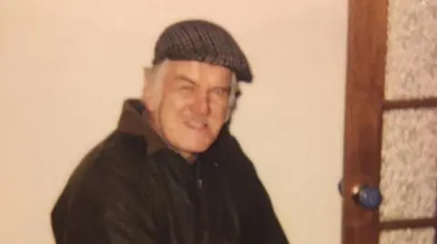 Family handout A faded photograph of an Michael Gwilliam. He is inside with a plain wall behind him and a wooden door to his right. He's wearing a wax jacket and a flat cap.