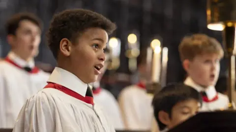A young chorister sings in a chapel. Behind him are other boys and men. All are wearing white cassocks with red collars.
