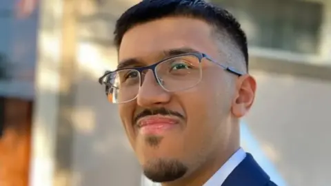 Family photo Head and shoulders photo of Sachin Hawkins. He has short black hair, a short beard and mustache, and smiles with his mouth closed. He wears half-rimmed glasses, a blue jacket and a white shirt.