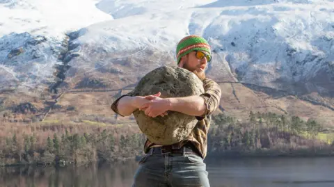 Calum Stott Calum Stott stands in front of snow-covered mountains and a tarn. Stott is lifting a granite-looking ancient stone. He has his mouth open in what looks like he is exerting himself. He is hugging the rock as he lifts it up. It is a sunny, wintery day and he is wearing sunglasses and a brown top and a green and orange hat.