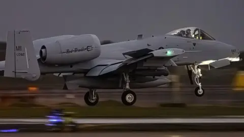Grey coloured jet aircraft. The pilot can be seen in the cockpit. The wheels are down as it comes in to land. The background is blurred owing to the motion of the aircraft. The picture was taken at night.