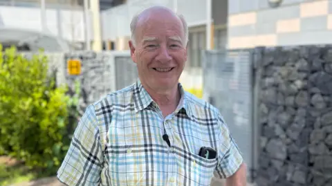 Deputy George Oswald - A man in his 60s with grey hair which is bald on the top smiling at the camera. he is wearing a short sleeve shirt which has a black, blue and yellow gingham pattern.