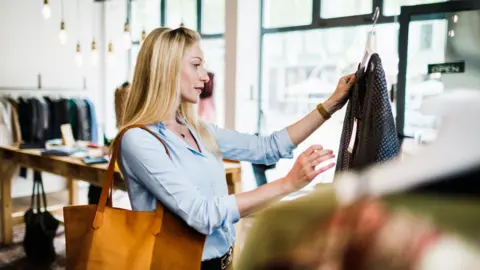 A woman with long blond hair looks at a top in a clothes shop. The woman is wearing a light blue top and has a light brown bag on her shoulder