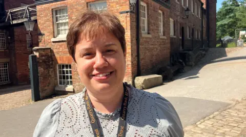 Photograph of Roz Stone, general manager of Quarry Bank Mill in Styal, Cheshire. She has short brown hair and is smiling for the camera. Wearing a patterned light grey top and with a National Trust lanyard around her neck, she is standing in front of the former cotton mill in the middle of the estate.