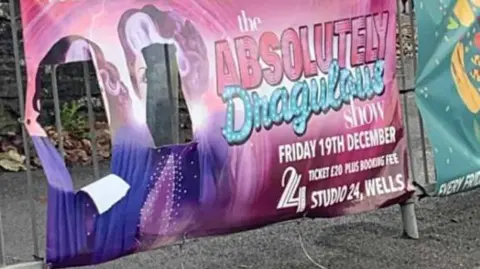 Facebook A photo of the defaced poster, you can read the writing but there are two holes cut out where the artists faces should be. It is clearly a poster attached to railings on a pavement. 