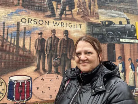 Tracey in front of the mural on Albion Street, with the words Orson Wright behind her above three workers 