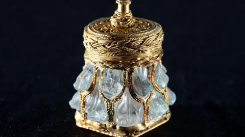 Neil Hanna A rock crystal jar, which resembles an ornate perfume bottle decorated in gold embellishments on a black background.