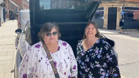 Two women wearing flowery tops propped against the back of a car with the boot open in an on-street bay in a street in a town centre with red-brick buildings to either side. The woman on the left has wavy dark hair and wears sunglasses, a white top with purple pattern and a dark shoulder strap. The woman on the right has dark, chin-length hair and wears a black top with pink and blue pattern.