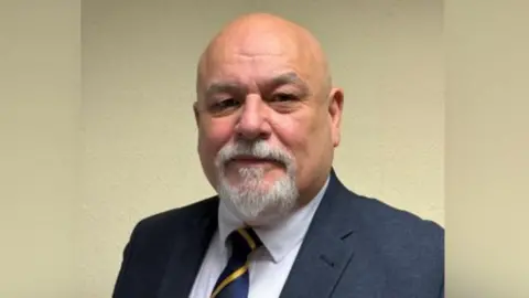 John Roddy - A bald middle aged white man with a grey goatee beard. He wears a navy suit and striped tie 