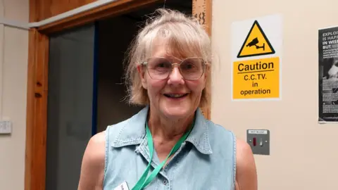 Jo Treeby smiling at the camera, wearing a denim sleeveless top, in the background is the custody reception where there is a wooden measuring stick.